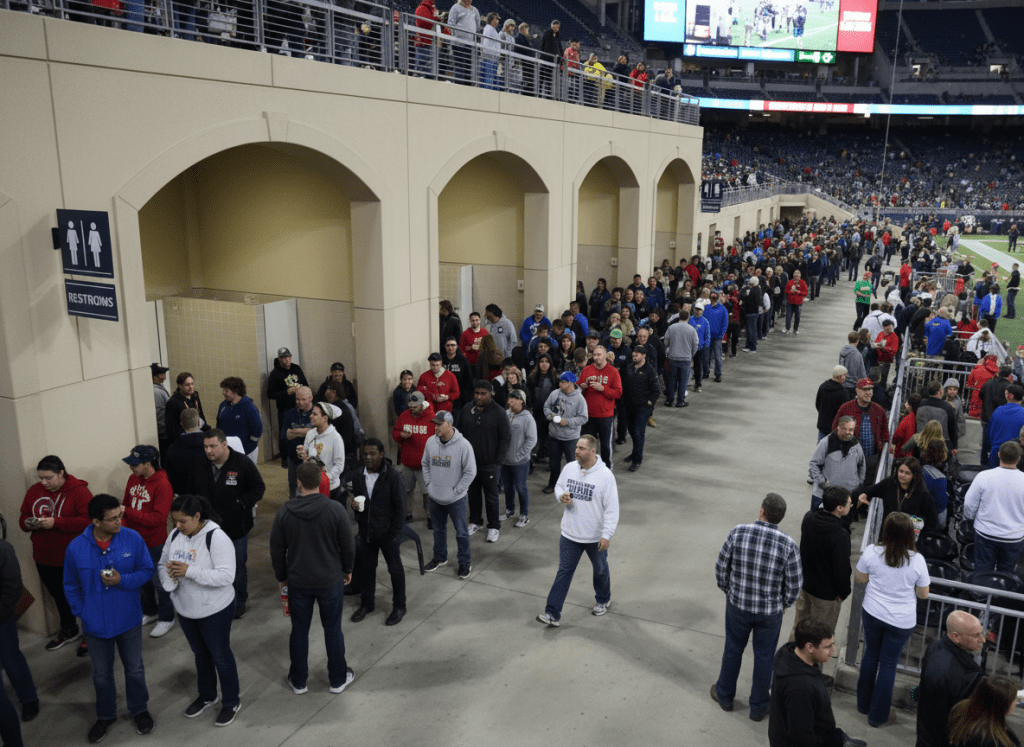 Long restroom line at a stadium or large event venue, diverse crowd, no visible brands or logos.